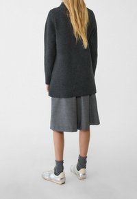 Langer Strickmantel in Anthrazit, grauer Plisseerock, silberne Sportschuhe und graue Socken. Fein gestrickte Textur, lockere Passform.