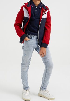 Niño con vaqueros azul claro, zapatillas blancas, polo azul marino y chaqueta con cremallera roja, blanca y azul marino, de pie con una mano en el bolsillo.