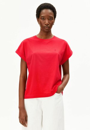 Femme aux cheveux courts et bouclés portant de petites boucles d’oreilles anneaux, un haut rouge à manches courtes et un pantalon blanc, une main dans la poche, expression neutre.