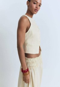 Beiger, kurz geschnittener Top mit Knopfleiste und breiten Trägern, kombiniert mit einem gerafften, cremfarbenen Rock. Das Model trägt bunte Harzarmbänder an einem Arm.