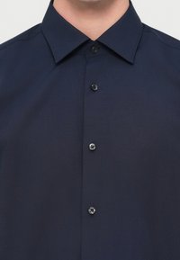 Πουκάμισο μπλε ναυτικό, με κουμπιά, κατασκευασμένο από ομαλό ύφασμα, με κλασικό γιακά και μαύρα κουμπιά κατά μήκος του μπροστινού μέρους.