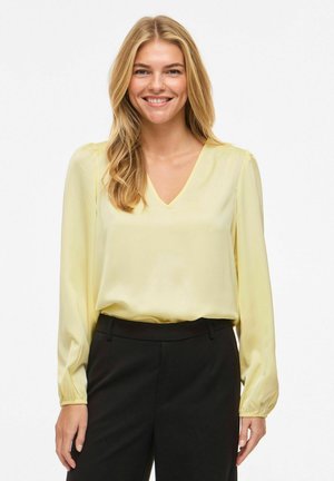 Blouse en soie jaune avec un col en V, manches longues avec poignets froncés, et une coupe ample. Associée à un pantalon tailleur noir.