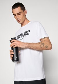 Hvit t-skjorte med "SEIER STARTER MED TRENING" trykket i svart. Person som holder en svart Nike vannflaske, med tatoveringer synlig på armen.