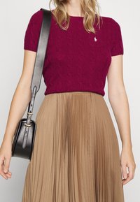 Fuchsia stickad topp med korta ärmar och kabelmönster, kombinerad med en plisserad beige kjol. Svart väska med gulddetaljer på axeln.