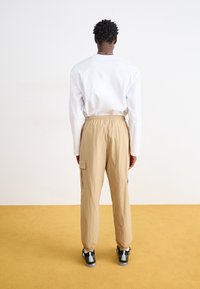 Beige cargo bukser med elastisk talje og håndled, med sidelommer og et glat, let væv. Båret med en hvid langærmet skjorte.