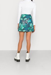 Kvinna klädd i en vit långärmad topp, grön blommig minikjol och vita snörboots som står vänd bort från kameran mot en enfärgad bakgrund.