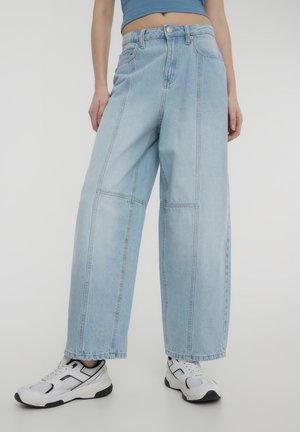 Jeans a gamba larga di colore blu chiaro realizzati in denim con cuciture a contrasto. Presentano una vita alta e tasche, abbinati a scarpe sportive bianche.