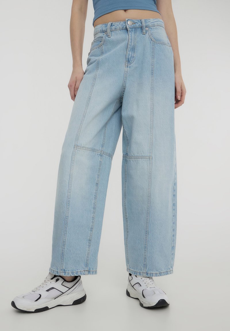 Jasnoniebieskie szerokie dżinsy wykonane z denimu z kontrastowymi przeszyciami. Posiadają wysoki stan i kieszenie, noszone z białymi butami sportowymi.