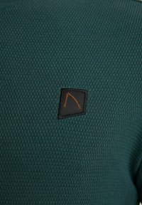 Tissu texturé vert foncé avec un petit patch noir en forme de losange présentant un motif abstrait de lignes orange cousu au centre.