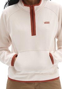 Suéter de color beige claro con un cuello de media cremallera y acentos naranjas. Cuenta con un bolsillo frontal tipo canguro y una tela suave y texturizada. Logo de la marca visible.