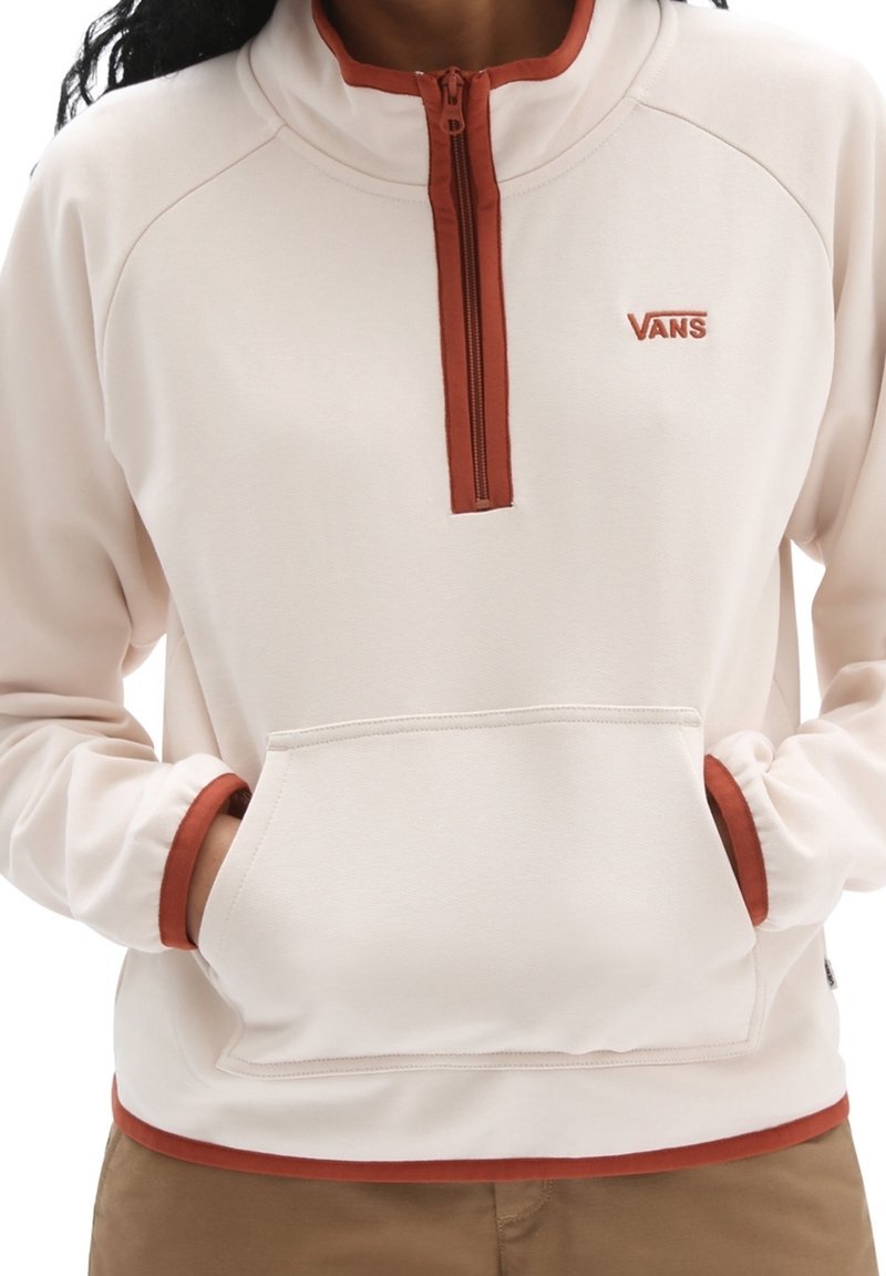 Suéter de color beige claro con un cuello de media cremallera y acentos naranjas. Cuenta con un bolsillo frontal tipo canguro y una tela suave y texturizada. Logo de la marca visible.