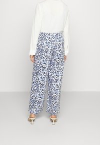 Woman wearing white long-sleeve top, wide-leg pants with blue and black leopard print, and white heeled sandals, standing against a plain background.