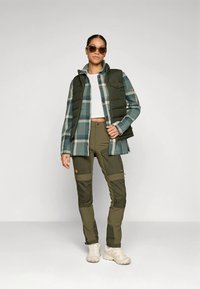 Πράσινο γιλέκο με επένδυση πάνω από μπλε και λευκό καρό πουκάμισο, συνδυασμένο με παντελόνι ελιάς με αντίθετα πάνελ και μπεζ αθλητικά παπούτσια.