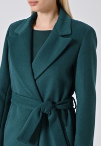 Teal wikkeljas van een textiel met structuur, grote revers en een ceintuur om de taille, met lange mouwen en een getailleerd ontwerp.