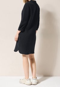 Schwarzes, strukturiertes Hemdkleid mit Kragen und 3/4-Ärmeln, kombiniert mit hellen Sneakers; mittellange, schulterlange Schnitte an den Seiten.