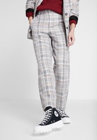 Szare spodnie w kratę o dopasowanym kroju, z kieszeniami i gładką fakturą. W połączeniu z czarnymi sneakersami typu high-top i białą gumową podeszwą.