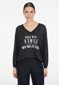 Schwarzes, leichtes Langarmoberteil mit V-Ausschnitt und glitzernder Textur. Mit weißem Drucktext: "REBEL WITH A SMILE" und locker sitzend.