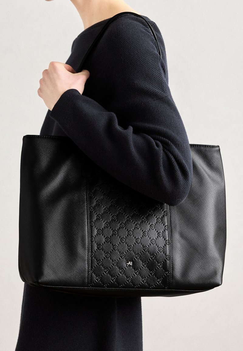 Personne portant un haut noir à manches longues tient un grand sac cabas en cuir noir avec un panneau central texturé et un petit logo argenté.