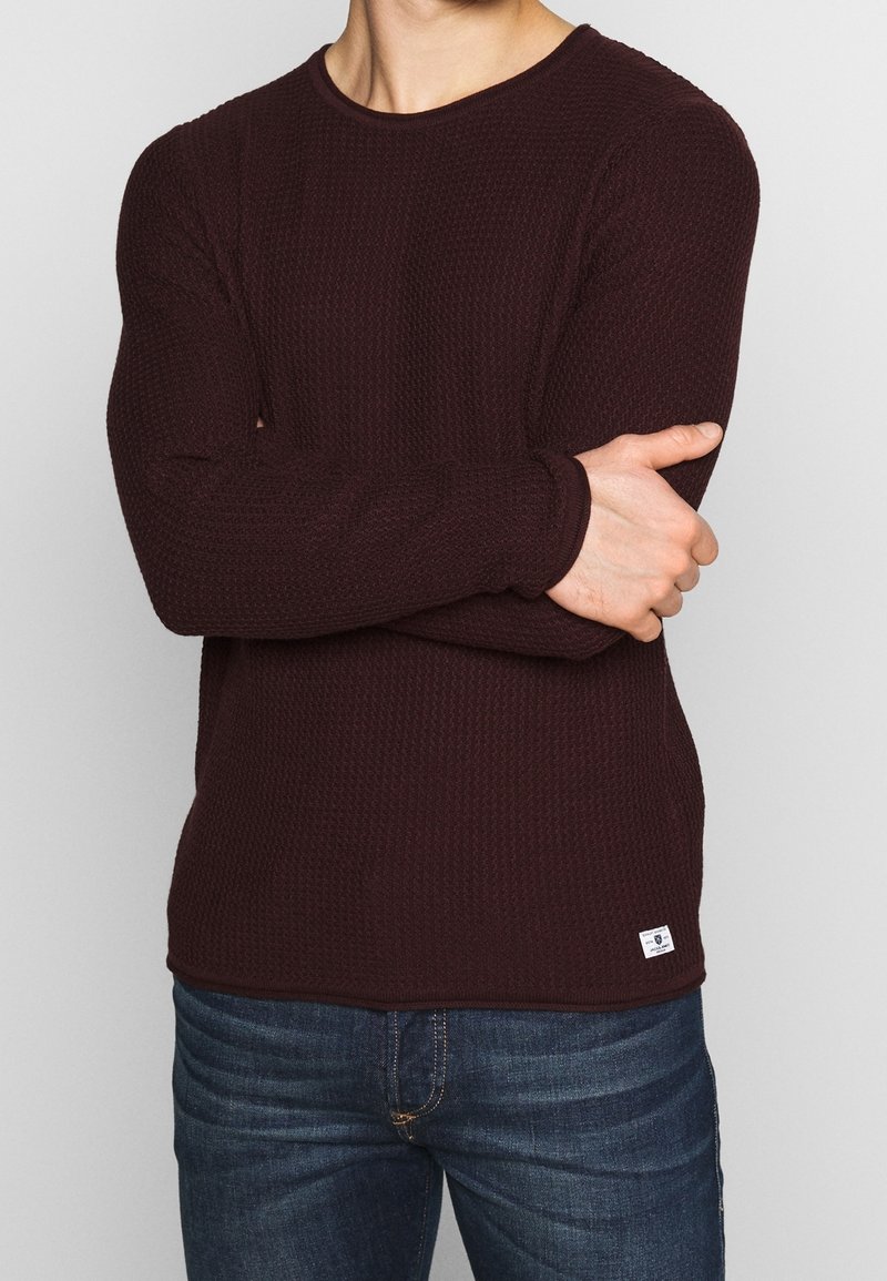 Bordowa koszula z długim rękawem o teksturowanym wzorze dzianiny i okrągłym dekolcie, zestawiona z niebieskimi dżinsami. Mała metka z logo na dole.