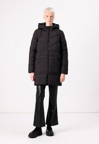 Manteau en duvet noir avec capuche, présentant un matelassage en chevrons, une fermeture éclair à l'avant et des poches latérales. Porté avec un pantalon noir évasé.