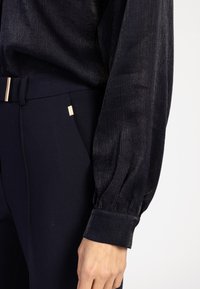 Zwarte gebloemde blouse met lange, ballonvormige mouwen, gecombineerd met donkere recht gesneden broeken met een subtiele glans en details in goudtint.