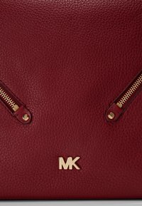 Gros plan sur un cuir rouge texturé avec deux fermetures éclair dorées et un logo MK doré sur un sac à main.
