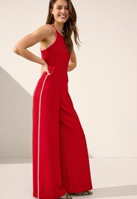 Röd ärmlös jumpsuit med sidostripes, vida ben och en figursydd bodice. Slät textur och kontrasterande kant accentuerar dess design.