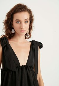 Schwarzes Kleid mit tiefem V-Ausschnitt, strukturiertem Stoff und gerafftem Volant an der Taille. Mit Schleifenakzenten an den Schulterträgern.