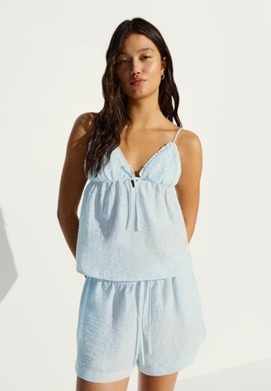 Mlada žena s dugom tamnom kosom, odjevena u svijetloplavu majicu bez rukava i pripadajuće kratke hlače, stoji na jednobojnoj bijeloj pozadini.