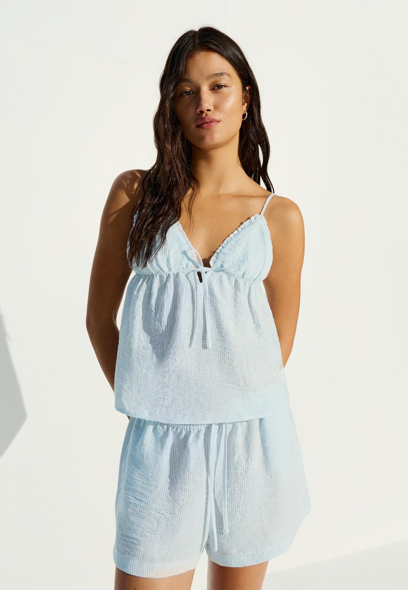Jeune femme aux longs cheveux noirs portant un débardeur bleu clair et un short assorti, debout devant un fond blanc uni.