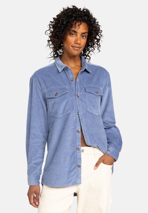 Blå corduroy skjorte med lange ærmer, to brystlommer, knaplukning og struktureret ribbet overflade; model iført off-white bukser.
