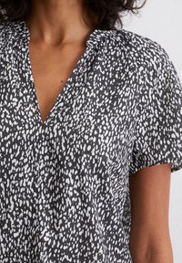 Short-sleeve blouse in black with white abstract spots, featuring a V-neck, soft fabric, and gathered detail at the shoulders.