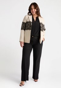 Blazer beige con detalles de encaje sobre una blusa negra, combinado con pantalones negros. El conjunto presenta un ajuste relajado y texturas elegantes.