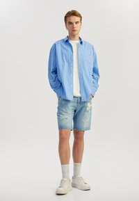 Chemise boutonnée à rayures bleu clair sur un t-shirt blanc, associée à un short en denim déchiré et des baskets blanches avec des semelles texturées.