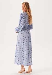 Lichtblauwe jurk met bloemenprint, lange pofmouwen, gerimpelde taille, enkel-lang, gemaakt van zachte stof. Draagt met doorzichtige hakken.