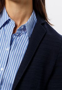 Blazer en maille bleu marine porté sur une chemise boutonnée bleu clair à rayures. Le blazer présente un tissu texturé, tandis que la chemise a des rayures blanches verticales.