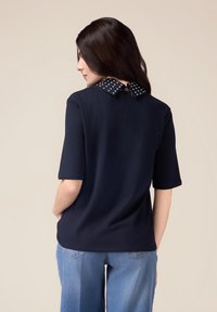 Haut à manches courtes bleu marine avec un col à pois, doté d'une fermeture à nouer dans le dos, associé à un jean bleu clair. Texture de tissu lisse.