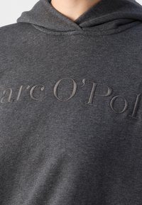Šedá mikina s kapucí s texturovaným, vyšitým logem "Marc O'Polo" v odpovídající barvě. Materiál je měkký a má jednoduchý, ležérní design.
