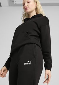 Schwarzes Hoodie- und Jogginghosen-Set aus weichem Stoff, mit einer Kängurutasche und dem weißen Puma-Logo am linken Hosenbein.