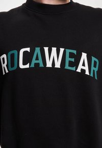 Musta huppari, jossa on ribattu kaula-aukko ja jossa rinnan kohdalla on sana "ROCAWEAR" rohkeasti valkoisella ja teal-värisellä tekstillä.