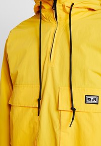Yellow pullover jacket with a half-zip design, two front pockets, adjustable hood, and black drawstrings. Features a small logo patch.