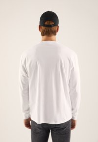 Weißes Langarmhemd mit Rundhalsausschnitt, getragen mit dunklen Jeans und einer schwarzen Baseballmütze, von hinten betrachtet.