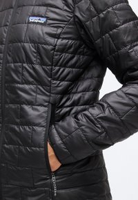 Person trägt eine schwarze gesteppte Patagonia-Jacke mit der Hand in der seitlichen Reißverschlusstasche, sichtbares Markenlogo auf der Brust.