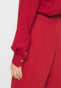 Blusa roja texturizada de mangas largas y puños abotonados; primer plano que muestra una tela suave y detalles de botones dorados brillantes.