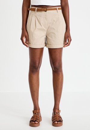 Persoon die beige shorts met hoge taille draagt, voorzien van een gevlochten bruine riem en sandalen met bruine bandjes, staand tegen een effen witte achtergrond.