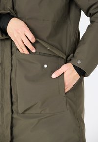 Olivgrön jacka med sidoficka och snapsförslutning, tillverkad av ett slätt, hållbart material. Har en stängbar klaff och dolda sömmar.