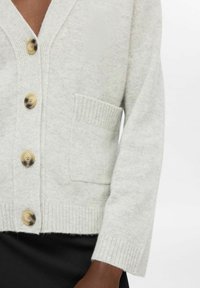 Šviesiai pilkas mezgimo kardiganas su V formos iškirpte, trimis dideliais mygtukais, raukšlėtomis rankovėmis ir kišene. Minkšta tekstūra ir kasdieniškas dizainas.