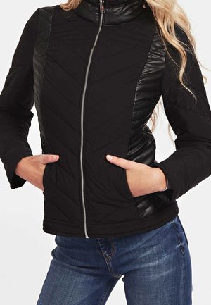 Femme portant une veste matelassée noire avec des détails en cuir, une fermeture éclair à l’avant et les mains dans les poches latérales, associée à un jean bleu.
