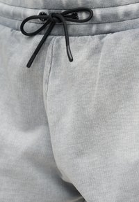 Ανοιχτό γκρι παντελόνι φόρμας με ριμπ ελαστική μέση και μαύρο κορδόνι δεμένο σε φιόγκο, που δείχνει την υφή και τις λεπτομέρειες της μπροστινής τσέπης.