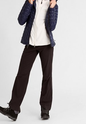 Osoba ubrana w czarne spodnie trekkingowe, czarne buty trekkingowe, białą kurtkę zapinaną na zamek oraz granatową pikowaną kurtkę, stojąca ze skrzyżowanymi nogami.